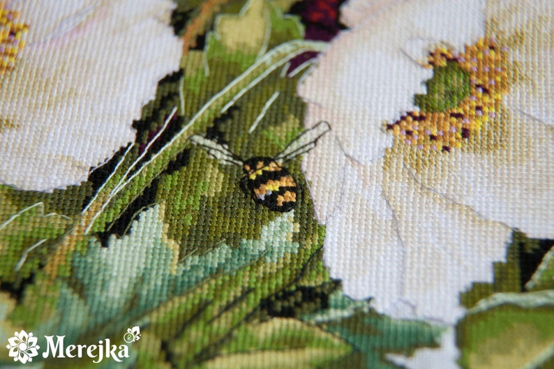 May include: Close-up of a cross-stitch embroidery depicting a white flower with a bee in the center. The bee is yellow and black with a brown body. The flower is surrounded by green leaves.