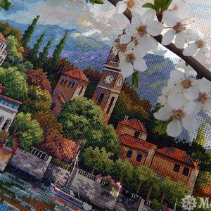 K&ouml;nnte beinhalten: Kreuzstich-Stickerei einer farbenfrohen Stadtlandschaft mit einem Uhrturm, Geb&auml;uden, B&auml;umen und einem Boot auf einem Fluss. Die Szene wird von wei&szlig;en Blumen eingerahmt.