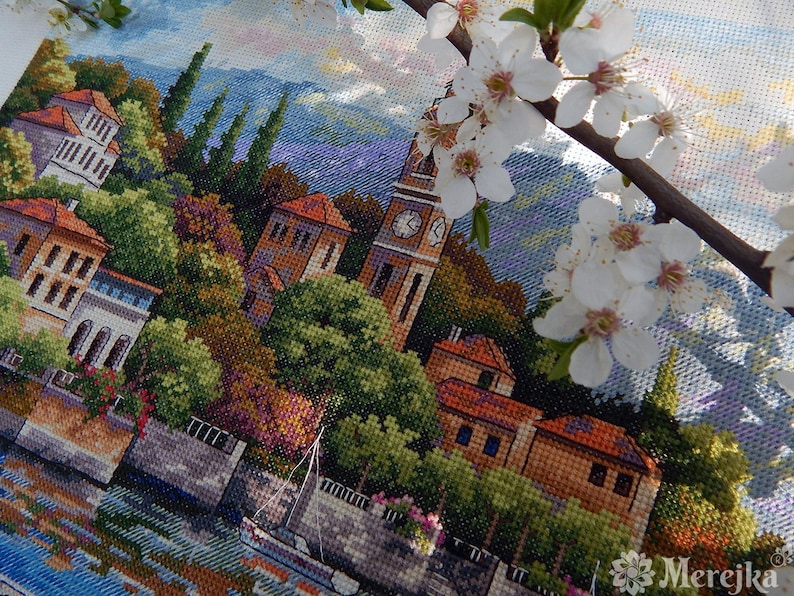 K&ouml;nnte beinhalten: Kreuzstich-Stickerei einer farbenfrohen Stadtlandschaft mit einem Uhrturm, Geb&auml;uden, B&auml;umen und einem Boot auf einem Fluss. Die Szene wird von wei&szlig;en Blumen eingerahmt.