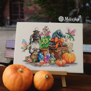 May include: A cross-stitch embroidery on canvas featuring a lantern, a potted plant, a blue hat with an orange bow, two pumpkins, glass potion bottles, and two winged frogs. The text Merejka is printed in the upper right corner of the canvas.
