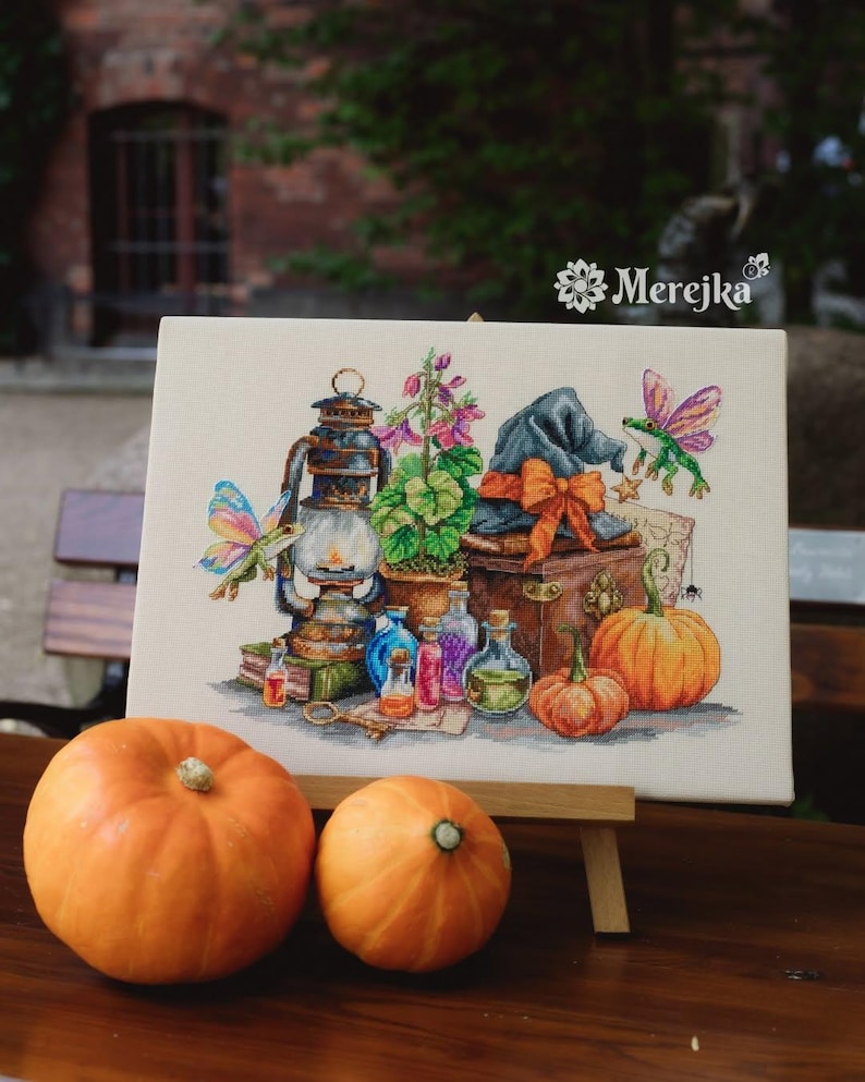May include: A cross-stitch embroidery on canvas featuring a lantern, a potted plant, a blue hat with an orange bow, two pumpkins, glass potion bottles, and two winged frogs. The text Merejka is printed in the upper right corner of the canvas.