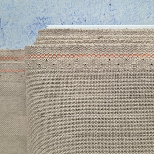 May include: Close-up of a beige linen fabric with a light orange stitching detail. The fabric is folded and rolled up.