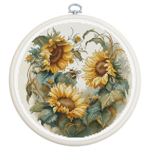 May include: A cross-stitch embroidery of sunflowers in a white hoop. The sunflowers are yellow and brown with green leaves and a bee. The embroidery is on a white background.