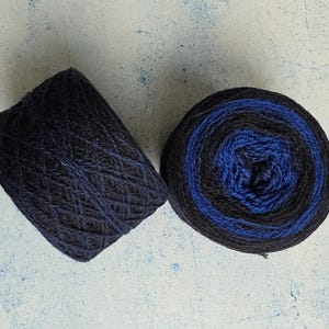 May include: Two skeins of yarn featuring a gradient color pattern that transitions from deep black to a vibrant royal blue. The yarn is wound into a compact, cylindrical shape, showcasing a textured, multi-ply fiber construction throughout.