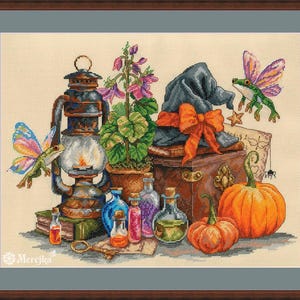 May include: A cross-stitch pattern featuring a dark lantern, a potted purple flower, a black hat with an orange bow, two pumpkins, and glass potion bottles. Two green frogs with colorful wings are present, along with a key and a small spider.