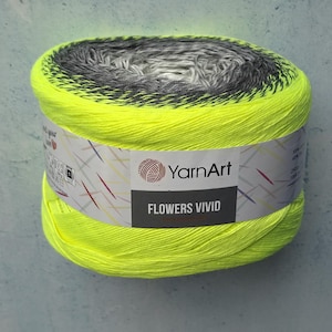 Może przedstawiać: Cylindryczny motek włóczki YarnArt Flowers Vivid o żywym neonowo-żółtym kolorze zewnętrznym, który przechodzi w ciemnoszary i biały środek. Biała papierowa etykieta owija środek, prezentując nazwę marki oraz linię produktu.