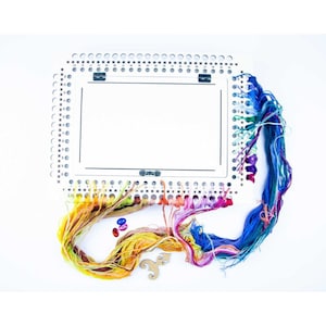 May include: A white plastic embroidery hoop with a white frame and a grid of holes. The hoop is surrounded by colorful embroidery floss, beads, and a wooden charm.