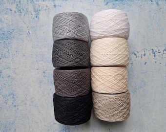 May include: Eight skeins of yarn in various neutral colors, including gray, white, black, and beige. The yarn is arranged in two columns of four skeins each, with a textured appearance. The background is a mottled blue and white surface.
