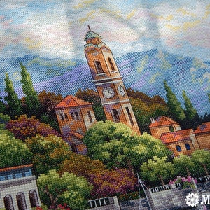 K&ouml;nnte beinhalten: Eine Kreuzstich-Stickerei, die ein malerisches italienisches Dorf mit einem Glockenturm zeigt, umgeben von &uuml;ppigem Gr&uuml;n und einer Bergkette im Hintergrund.