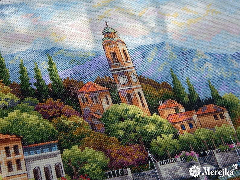K&ouml;nnte beinhalten: Eine Kreuzstich-Stickerei, die ein malerisches italienisches Dorf mit einem Glockenturm zeigt, umgeben von &uuml;ppigem Gr&uuml;n und einer Bergkette im Hintergrund.