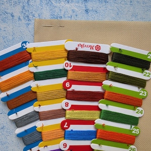 May include: A collection of embroidery floss organized on numbered cardboard bobbins, featuring a variety of colors including red, yellow, green, blue, and brown. The bobbins are arranged over a piece of beige Aida fabric for cross-stitch projects.