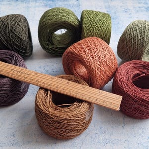 May include: A collection of colorful yarn skeins in shades of green, brown, orange, and purple. A wooden ruler rests across some of the skeins. The yarn is tightly wound, suggesting it's ready for crafting projects.