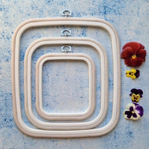 May include: Three nested rectangular embroidery hoops made of light-coloured wood, each featuring a silver metal hanging bracket at the top centre. The hoops are arranged from largest to smallest, with four colourful pansy flowers placed to the right.
