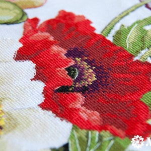 May include: Close-up of a red poppy flower with a purple center, surrounded by green leaves and white flowers. The fabric is a white Aida cloth, commonly used for cross-stitch embroidery.