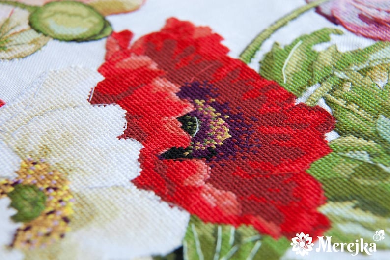 May include: Close-up of a red poppy flower with a purple center, surrounded by green leaves and white flowers. The fabric is a white Aida cloth, commonly used for cross-stitch embroidery.