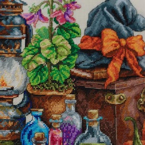 May include: A cross-stitch embroidery pattern featuring a brown lantern, a potted plant with pink flowers, a blue hat with an orange bow, and several glass bottles filled with colorful liquids. The text Merejka is printed in white at the top.