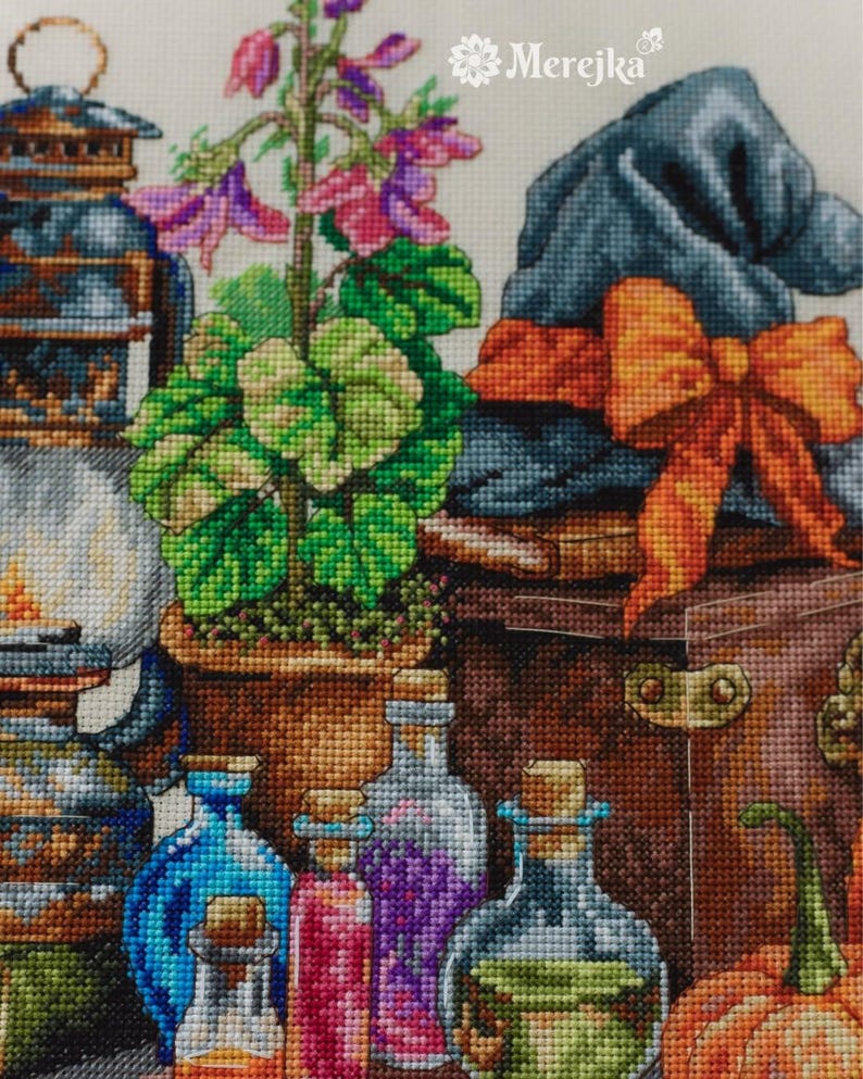 May include: A cross-stitch embroidery pattern featuring a brown lantern, a potted plant with pink flowers, a blue hat with an orange bow, and several glass bottles filled with colorful liquids. The text Merejka is printed in white at the top.