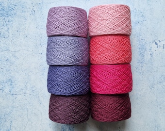 May include: Eight skeins of yarn in a variety of colors, including shades of pink, red, purple, and blue. The yarn is arranged in two columns of four, with each skein tightly wound. The background is a light blue and white textured surface.