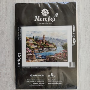 K&ouml;nnte beinhalten: Kreuzstich-Set mit einem farbenfrohen Bild einer Seestadt mit einem Kirchturm, betitelt "Lago di Como". Das Set enth&auml;lt Zweigart Aida-Stoff, DMC-Garn, eine Nadel und ein Symbolmuster. Das Set ist in einer schwarzen Plastikt&uuml;te mit dem Merejka-Logo und der Website-Adresse verpackt.