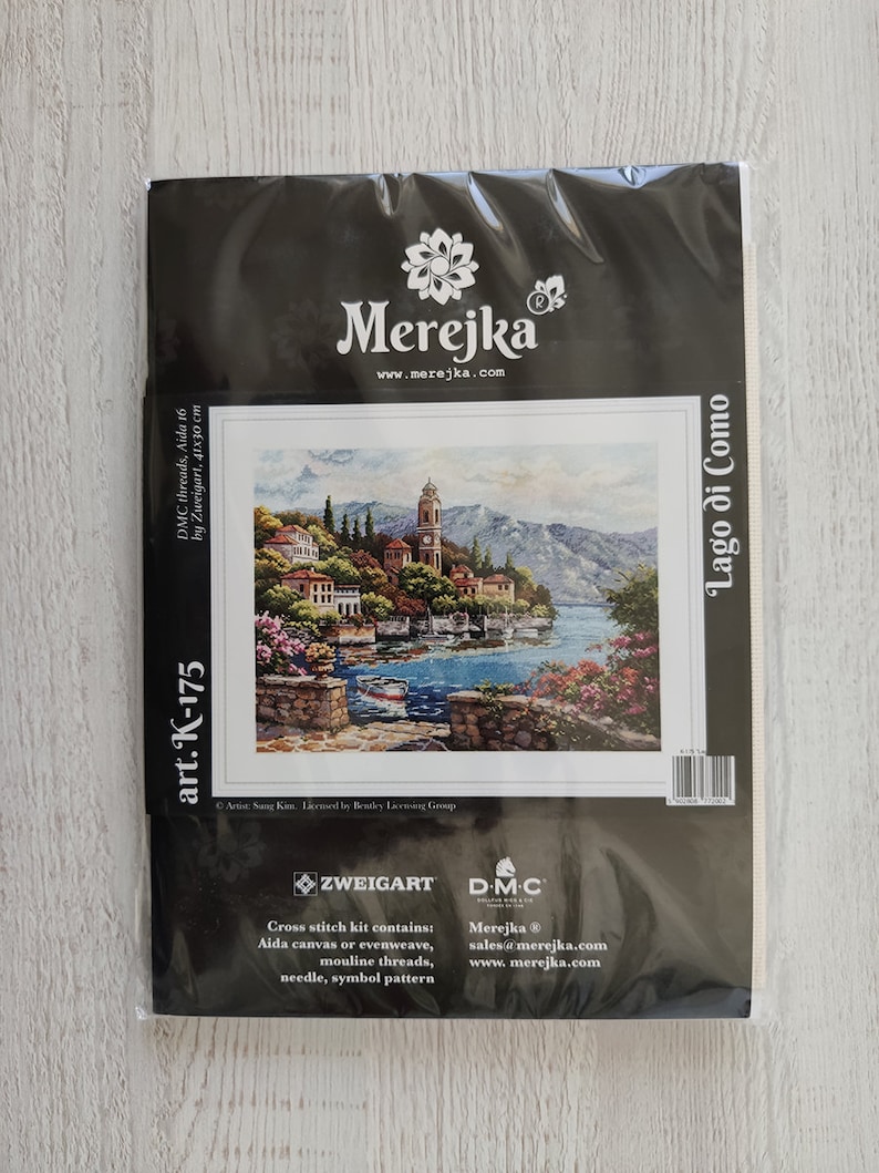 K&ouml;nnte beinhalten: Kreuzstich-Set mit einem farbenfrohen Bild einer Seestadt mit einem Kirchturm, betitelt "Lago di Como". Das Set enth&auml;lt Zweigart Aida-Stoff, DMC-Garn, eine Nadel und ein Symbolmuster. Das Set ist in einer schwarzen Plastikt&uuml;te mit dem Merejka-Logo und der Website-Adresse verpackt.