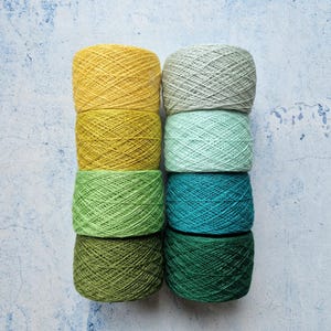 May include: Eight balls of yarn in shades of yellow, green, and teal, arranged in two columns. The yarn is displayed against a textured, pale blue background. Each ball is tightly wound.