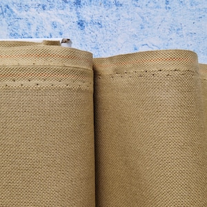 Puede incluir: Dos rollos de tela beige con una textura tejida sutil. La tela tiene una fina franja naranja a lo largo del borde y está cosida con pequeños puntos negros.