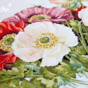 May include: Close-up of a cross-stitch embroidery featuring a bouquet of red, white, and pink poppies with green leaves. The embroidery is on a white fabric background.