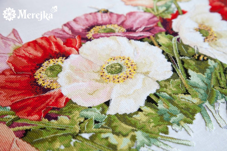 May include: Close-up of a cross-stitch embroidery featuring a bouquet of red, white, and pink poppies with green leaves. The embroidery is on a white fabric background.