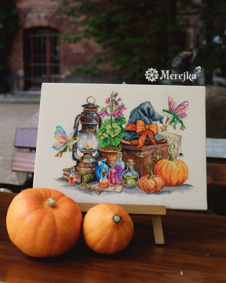 May include: A cross-stitch embroidery on canvas featuring a lantern, a potted plant, a blue hat with an orange bow, two pumpkins, glass potion bottles, and two winged frogs. The text Merejka is printed in the upper right corner of the canvas.