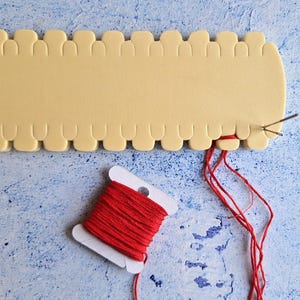 May include: A close-up of a beige foam loom with red thread threaded through the holes. A spool of red thread is on a white plastic spool holder. Two needles are also visible.
