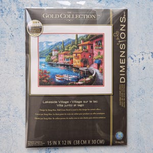 Può includere: Un kit a punto croce contato Dimensions Gold Collection con una scena di un villaggio sul lago. L'immagine mostra edifici colorati, barche e acqua. Il kit misura 38 cm x 30 cm.