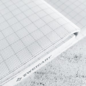 May include: Close-up of white Aida cloth with a grid pattern. The fabric is folded over and the edge shows the brand name "ZWEIGART" printed on the side.