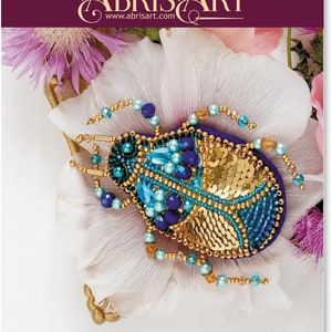 May include: A bead embroidery kit featuring a detailed, jewel-toned beetle design. The beetle is adorned with blue, turquoise, and gold beads, set against a white background with floral accents. The kit includes the text "ABRIS ART" and "Bead embroidery kit".