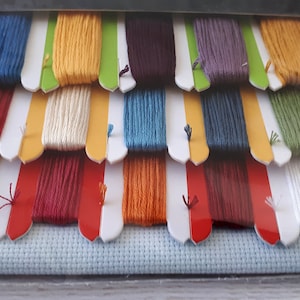 Puede incluir: Un conjunto de hilo de bordar en varios colores, incluyendo rojo, amarillo, azul, verde y blanco. El hilo est&aacute; organizado en filas y est&aacute; empaquetado en una bolsa de pl&aacute;stico transparente. Un trozo de tela Aida blanca est&aacute; incluido en el paquete.