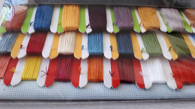 Puede incluir: Un conjunto de hilo de bordar en varios colores, incluyendo rojo, amarillo, azul, verde y blanco. El hilo est&aacute; organizado en filas y est&aacute; empaquetado en una bolsa de pl&aacute;stico transparente. Un trozo de tela Aida blanca est&aacute; incluido en el paquete.