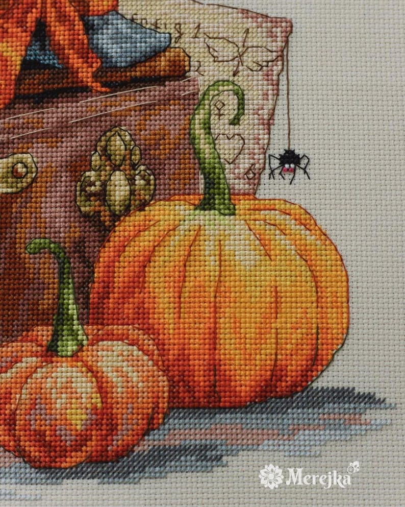 May include: A cross-stitch embroidery design featuring two orange pumpkins with green stems, a small black spider hanging from a thread, and a textured brown background. The word Merejka is printed in white in the bottom right corner of the fabric.