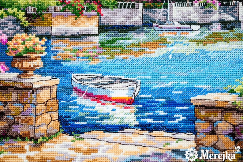K&ouml;nnte beinhalten: Eine Kreuzstich-Stickerei, die ein kleines Boot zeigt, das in einem blauen Gew&auml;sser angedockt ist. Das Boot ist wei&szlig; mit einem roten und gelben Streifen. Die Szene wird von Steinmauern und &uuml;ppigem Gr&uuml;n eingerahmt.