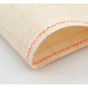 May include: Close-up of a folded, off-white cloth with a woven texture. The edge is finished with a bright orange stitch. The fabric appears to be a natural fiber, possibly linen or cotton, and is slightly translucent.