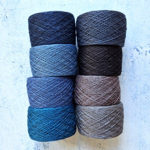 Puede incluir: Ocho madejas de hilo en varios tonos de azul, gris y marrón están apiladas en dos columnas. El hilo está bien enrollado y los colores varían del azul marino oscuro al gris claro y al marrón terroso. El fondo es una superficie texturizada blanquecina.