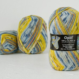 May include: Three balls of yarn in shades of blue, yellow and white. The yarn is striped and has a textured look. The label on one ball reads "Opal 100 g ca. 425 m 75% Schurwolle 25% Polyamid In Variation nach Hundertwassers Werk (05) Die vier Einsamkeiten."
