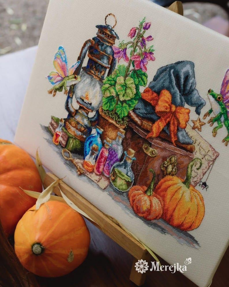 May include: A cross-stitch embroidery on fabric featuring a lantern, a potted plant, a dark hat with an orange bow, colorful potion bottles, two small winged creatures, and pumpkins. The design includes the text Merejka in the bottom corner.