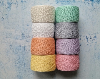 May include: An overhead shot displays eight skeins of yarn in a variety of colors. The colors include cream, mint green, light grey, dark grey, orange, lavender, yellow, and pink. The yarn is arranged in two columns of four skeins each, set against a blue and white textured background.