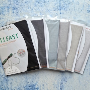 Peut inclure: Un paquet de tissu de lin Zweigart Belfast prédécoupé en différentes couleurs, dont le noir, le gris, le bleu et le beige. Le paquet comprend une étiquette avec le texte "Zweigart précoupé Belfast Premium Quality Made in Germany 126/cm 37 ct. 48x48cm 19x27" et une petite étiquette avec le texte "Bartpay ZORLAGE FREE CHART".