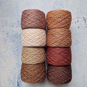 May include: Eight skeins of yarn in shades of brown, beige, and red-brown are stacked in two columns of four. The yarn is tightly wound, with colours ranging from light tan to deep chocolate. The background is a textured, light blue surface.