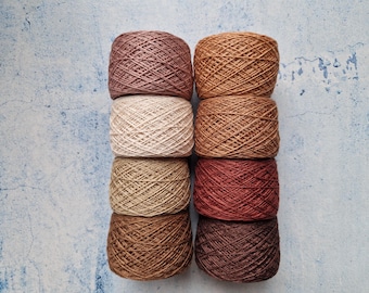 May include: Eight skeins of yarn in various shades of brown, beige, and red-brown are stacked in two columns of four. The yarn is tightly wound, and the colors range from light tan to deep chocolate. The background is a textured, light blue surface.