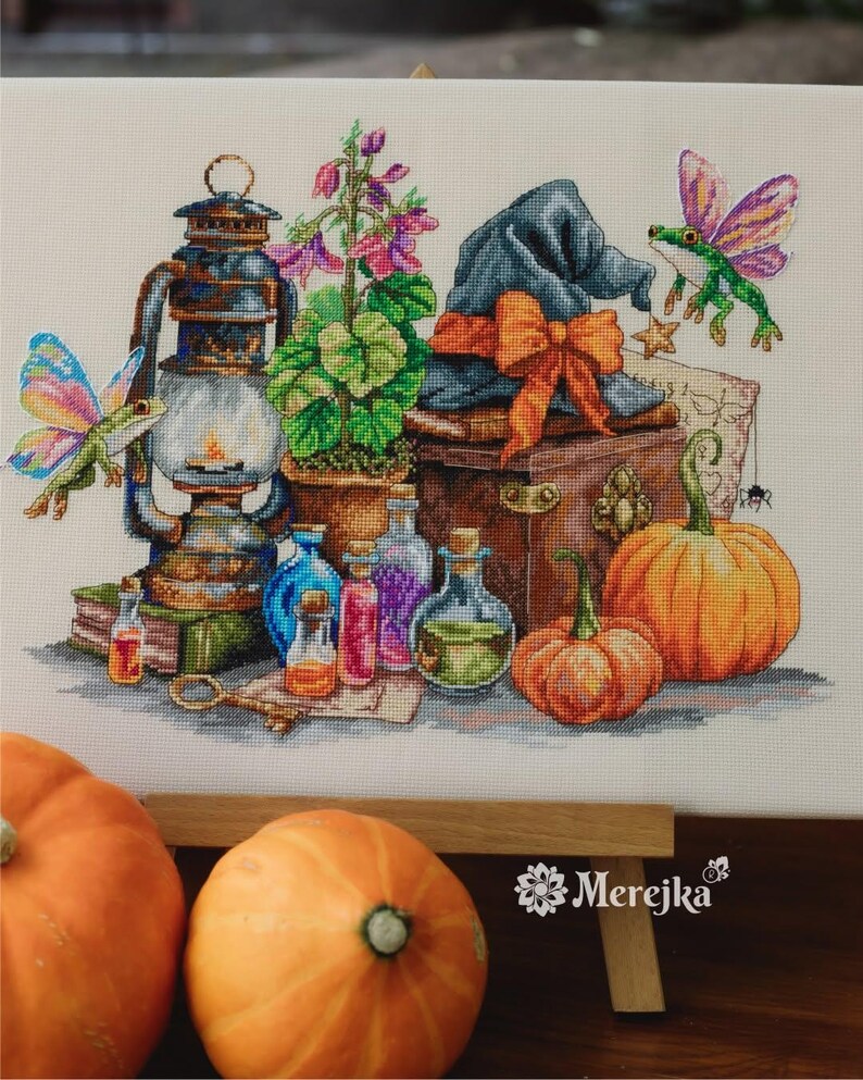May include: A cross-stitch embroidery featuring a lantern, a potted plant, a dark hat with an orange bow, two pumpkins, and two winged frogs. Several small glass bottles with colored liquids, a key, and a spider are also depicted in the scene.