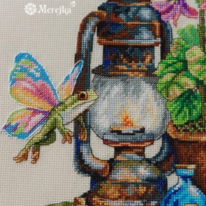 May include: A cross-stitch embroidery pattern featuring a green frog with multicolored wings next to a rustic metal lantern. The design includes a potted plant, a blue glass bottle, and a small clear bottle. The text Merejka is visible.