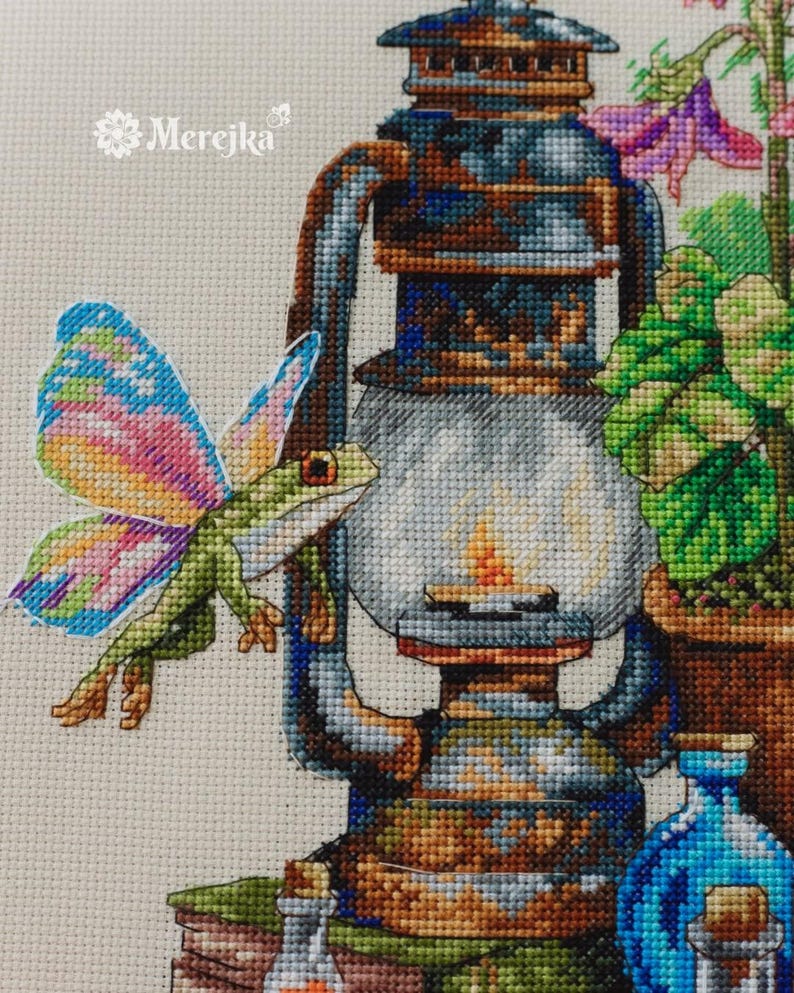 May include: A cross-stitch embroidery pattern featuring a green frog with multicolored wings next to a rustic metal lantern. The design includes a potted plant, a blue glass bottle, and a small clear bottle. The text Merejka is visible.