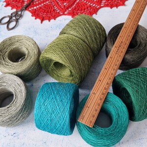 May include: An assortment of yarn skeins in various colors, including green, teal, and gray, are arranged on a light blue surface. A wooden ruler and a pair of scissors are also present. The yarn is likely for knitting or crochet projects.