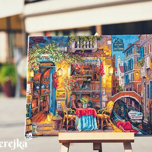 Puede incluir: Una obra de punto de cruz colorida que representa una pintoresca escena de calle italiana con un canal, un puente y un caf&eacute; con mesas y sillas. La obra est&aacute; enmarcada y expuesta en un caballete.